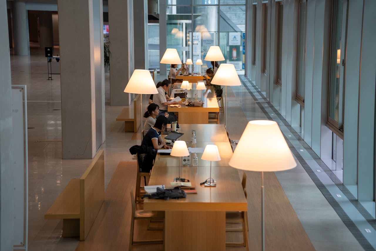 Spacious library interior with people studying at tables under warm lamps. Ideal for education or lifestyle themes.